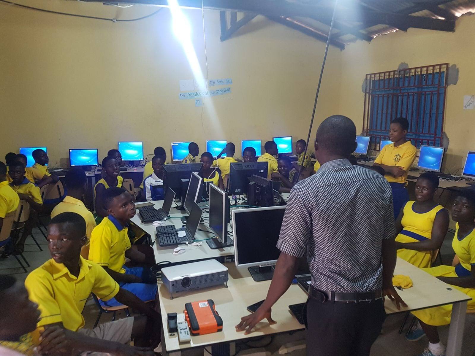 A computer classroom at a school in Ghana, with pupils in yellow and blue uniforms at rows of monitors while a teacher stands at the front bench.
